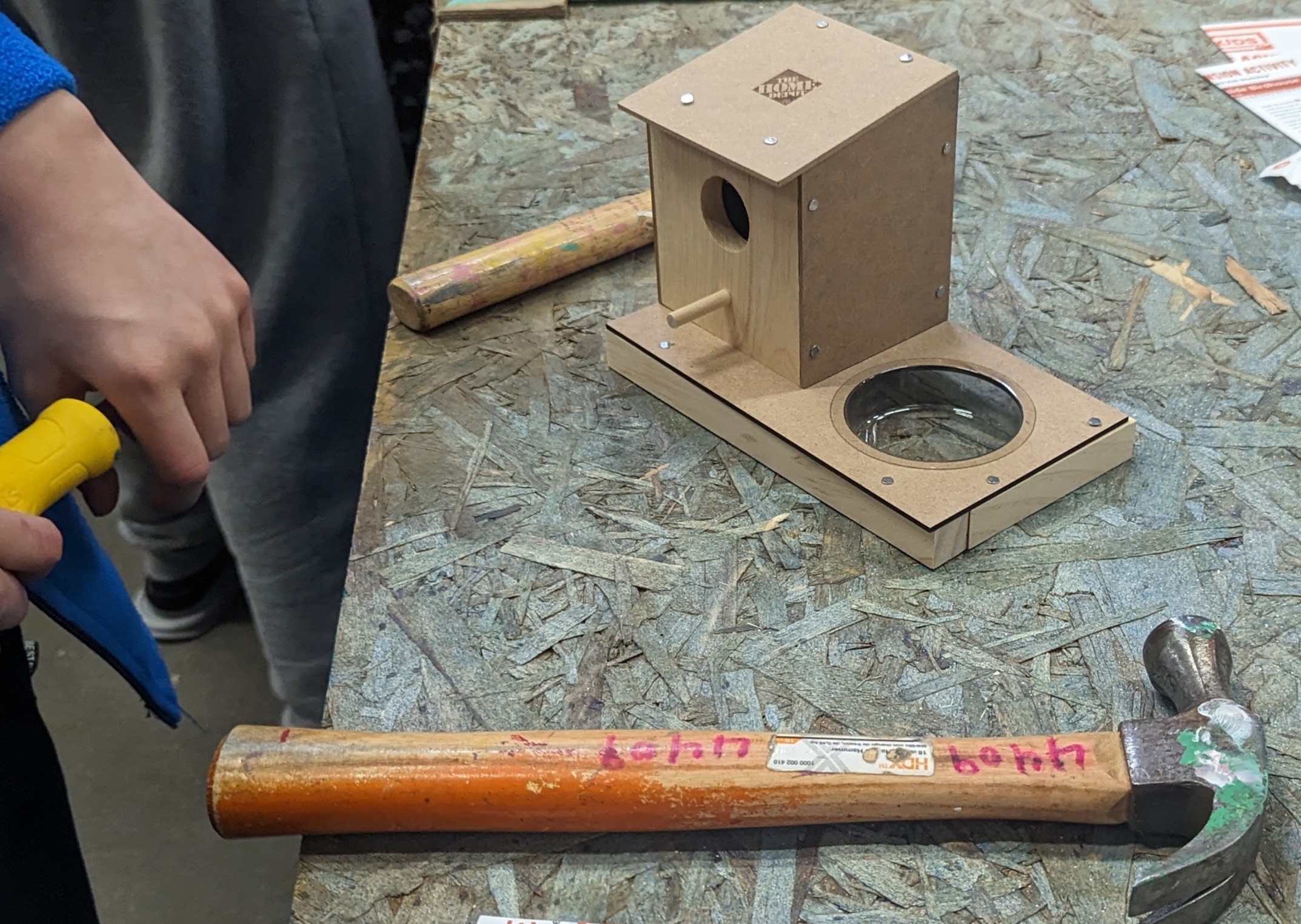 a birdhouse and hammer on a table with a child's hands nearby